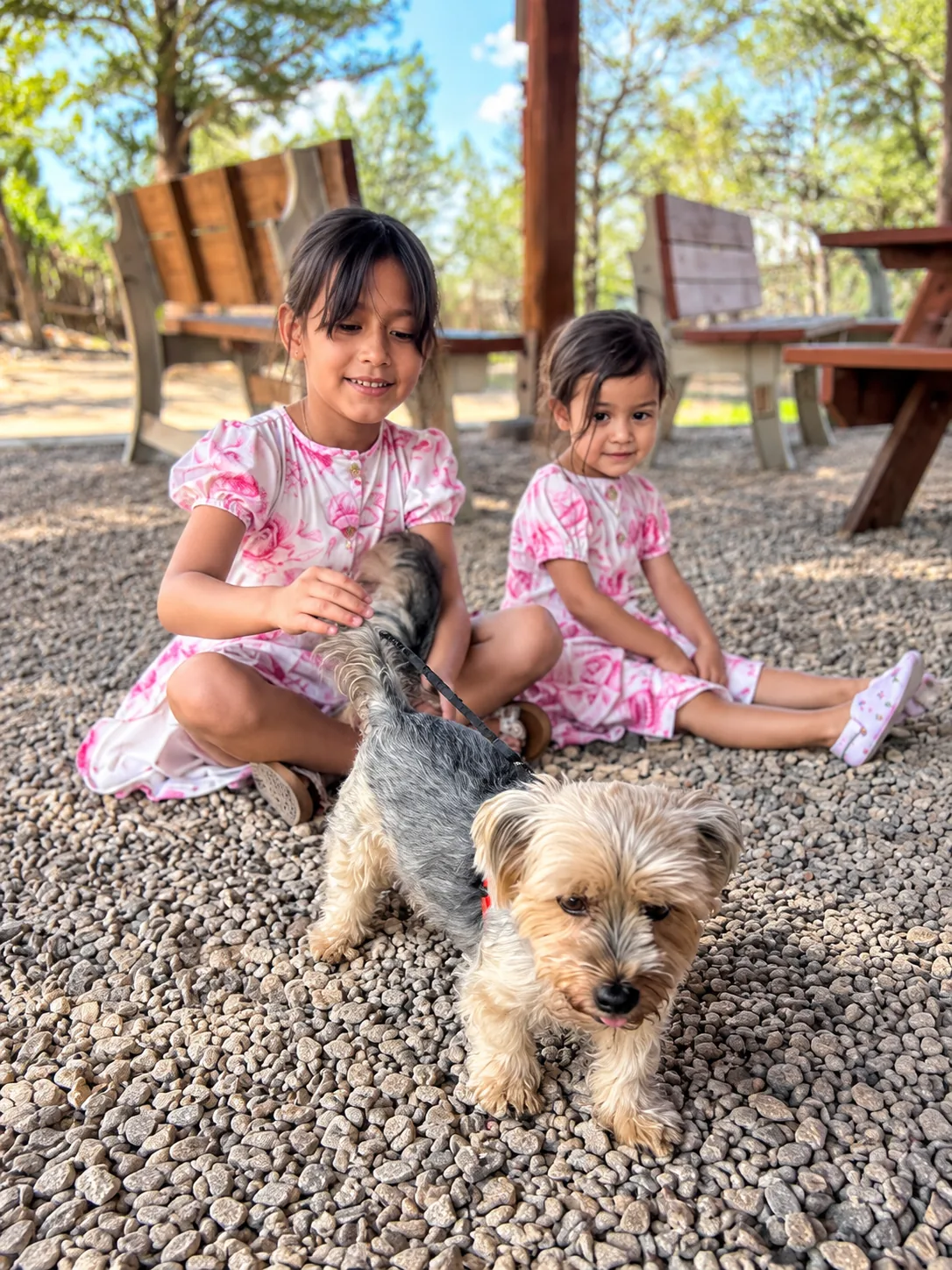 Two girls sitting on the pavilion gravel with a small dog on a leash